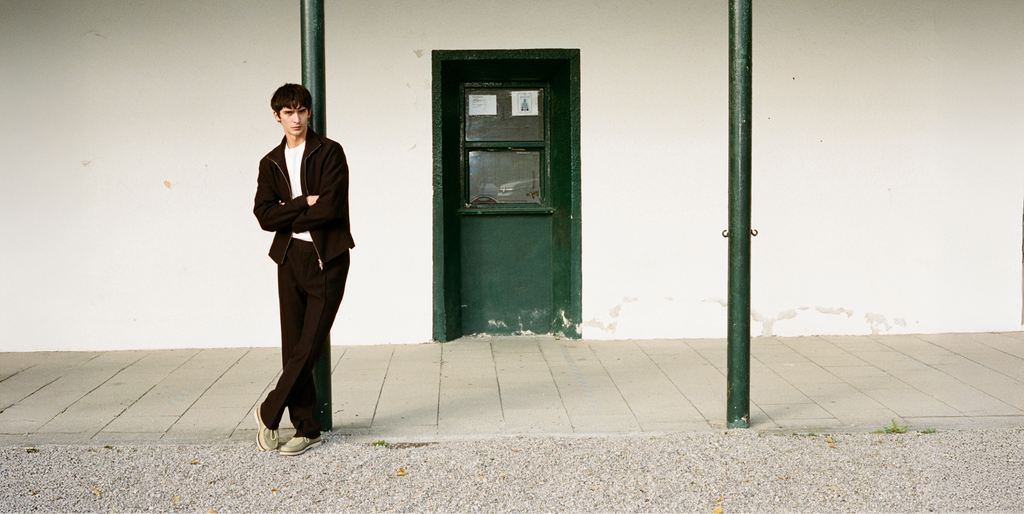 Brown haired man in a brown woolen tracksuit standing in front of a green door